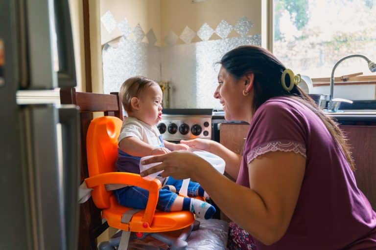 Latin american mother is feeding her baby with down syndrome in a highchair in their kitchen