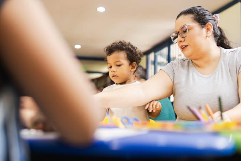 Teacher assisting a mixed race toddler boy with arts and crafts in a preschool classroom setting