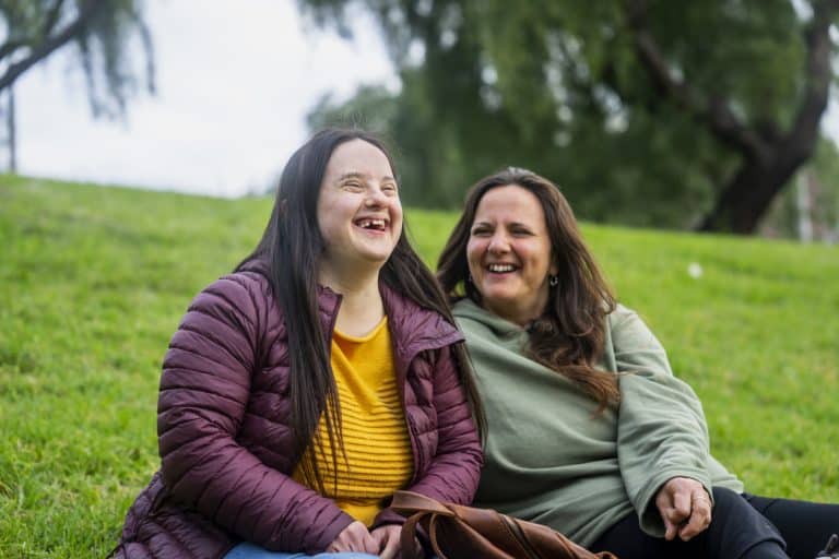 Portrait of woman with down syndrome laughing while sitting in grass with mother during daytime.