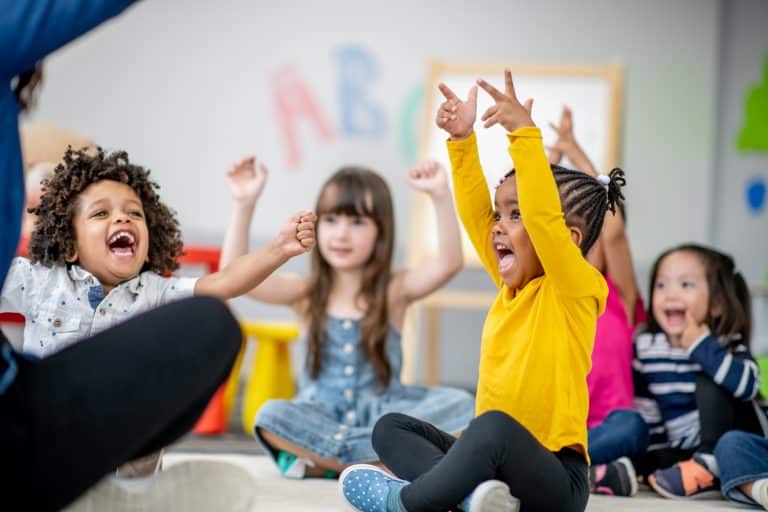 A small group of Kindergarten children sit on the floor of their classroom as their teacher lead them in a sing-along. They are each dressed comfortably and are smiling as they do the actions to the song.