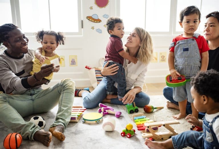 A diverse group of mothers and fathers play in a carpeted playroom with their children ranging from 2-4.