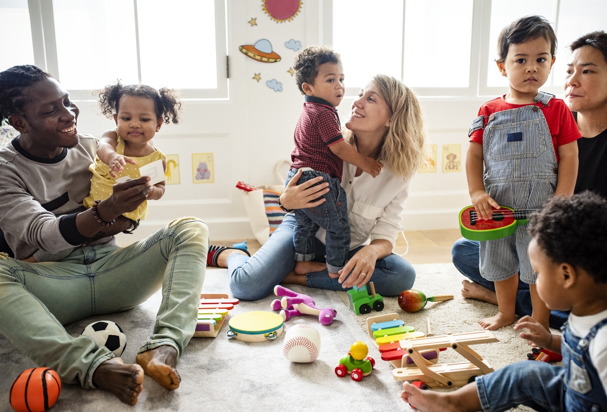 A diverse group of mothers and fathers play in a carpeted playroom with their children ranging from 2-4.