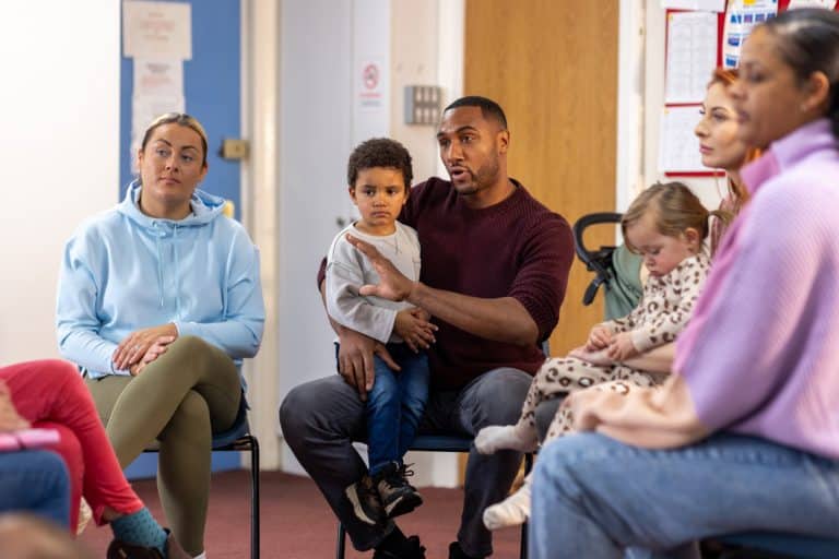 Over-the-shoulder shot of a father sitting down with his son on his lap. They are having a meeting with a childcare support worker out of shot and other parents in a community centre.