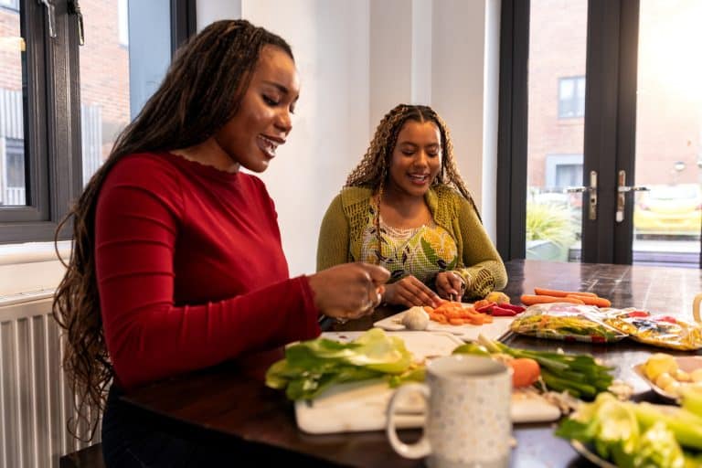 A medium shot of two university students, one of whom is neurodiverse, enjoying preparing a healthy meal together in their shared kitchen. The scene captures collaboration and inclusion, with both women smiling and engaged—one slicing carrots while the other arranges fresh vegetables. The warm atmosphere highlights friendship, independence, and the everyday life of diverse students in a supportive academic environment.