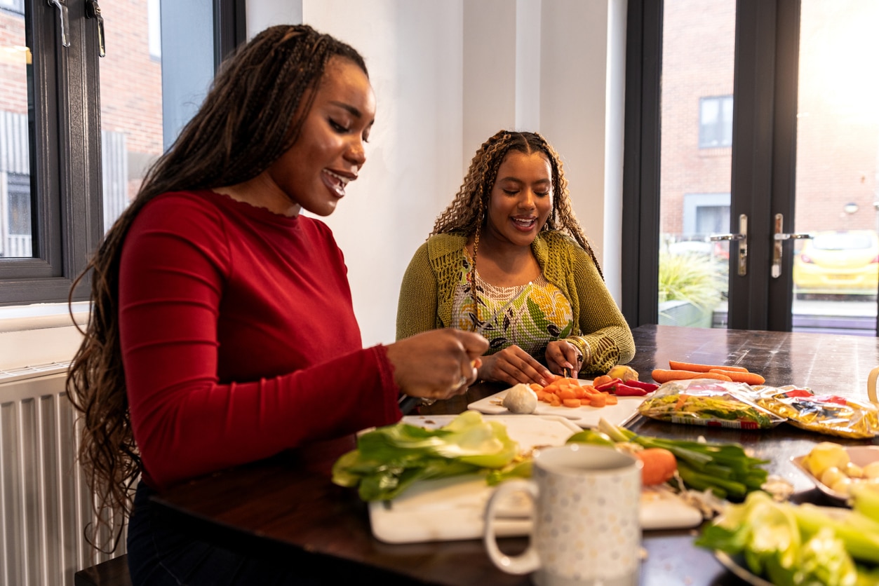A medium shot of two university students, one of whom is neurodiverse, enjoying preparing a healthy meal together in their shared kitchen. The scene captures collaboration and inclusion, with both women smiling and engaged—one slicing carrots while the other arranges fresh vegetables. The warm atmosphere highlights friendship, independence, and the everyday life of diverse students in a supportive academic environment.